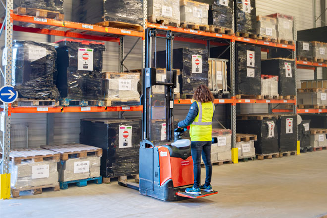 Apprenant utilisant un gerbeur électrique dans un entrepôt, portant un gilet haute visibilité devant des racks de stockage.